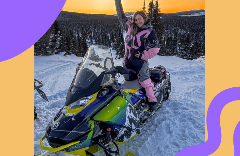 a picturesque orange sunset sky on top of a ice mountain with a super strong neon snowmobile. A women in pink ice gear raising her hands up high celebrating life and her success in the mining industry